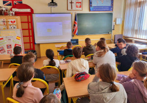 Wnętrze klasy.Na pierwszym planie widoczni uczniowie. Uczniowie siedzą w ławkach. Ujęcie klasy z tyłu. W tle widoczna tablica biała i tablica interaktywna z wyświetlonym obrazem.