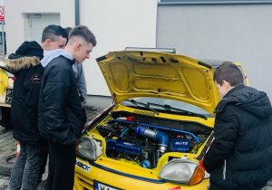 Żółty samochód z podniesioną maską. Przed samochodem stoi trzech chłopaków w ciemnych kurtkach. Chłopcy patrzą pod maskę.