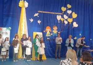 Dwie uczennice i kobieta stoją na niebieskim tle. Dziewczęta trzymają w dłoniach dyplomy i torby.
