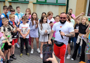 Okrąg utworzony z dużej grupy uczniów. W środku okręgu stoi mężczyzna w czerwonych spodniach i białej podkoszulce. Mężczyzna trzyma w ręku mikrofon. W tle budynek szkolny.