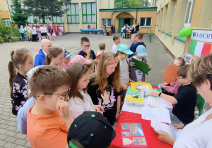 Stolik nakryty czerwonym płutnem.Przed stolikiem stoi duża grupa uczniów ubrana na kolorowo. W tle budynek szkolny.