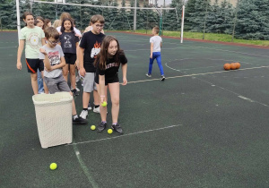 Boisko szkolne. Na boisku stoi grupa uczniów. Przed nimi stoi biały plastikowy kosz. Jeden z uczniów pochyla się . W ręku trzyma małą piłeczke.