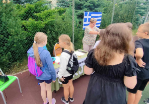 Boisko szkolne. Pod ogrodzeniem z siatki stoi stolik i trzy uczennice. Na siatce wisi flaga Grecji. Na pierwszym planie kobieta w czarnej sukience odwrócona plecami.