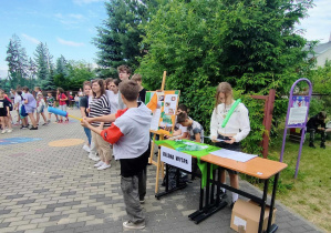 Wybieg szkolny. Na wybiegu znajdyje się duża grupa dzieci. Na pierwszym planie znajduje się stolik. Przy stoliku stoi dziewczyna z kartka w ręku.