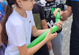 Na stoliku lezy pompka do balonów. Uczennica w białej bluzce trzyma w ręku długi zielony balon. Przy niej stoi kilku chłopców.