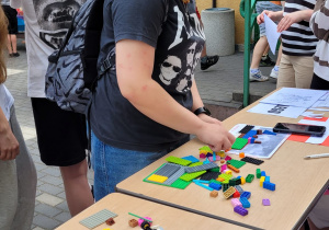 Dwa stoliki z klockami lego stojące po przekątnej kadru. Nad stolikiem pochykla się dziewczyna. Za nią stoi grupa uczniów i nauczycielka. W tle budynek szkoły.i