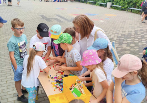 Wybieg szkolny. djęcie z góry .Widoczny stolik z klockami lego. Nad stolikiem pochyla się grupa uczniów. W górnej części zdjęcia stoi duża grupa uczniów. W tle drzewa.