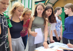 Grupa uczniów stoi przy stoliku z kartkami . W tle szkoła.