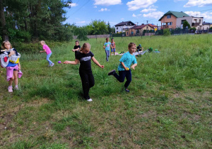 Grupka dzieci biegnie z łyżeczkami na których sa kolorowe piłeczki. Dzieci znajdują się na łące. W tle domki jednorodzinne.