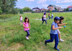 Grupka dzieci biegnie z łyżeczkami na których sa kolorowe piłeczki. Dzieci znajdują się na łące. W tle domki jednorodzinne.