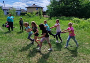 Grupka dzieci bawi się na łące. W tle domki jednorodzinne.