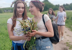 Dwie dziewczyny w wiankach i bukietem polnych kwiatów idą piaszczystą drogą. W tle inne dziewczęta, łaka i drzewa.