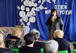 Pani dyrektor Ewa Brzezińska składa życzenia wszystkim pracownikom szkoły. Za panią dyrektor widać dekorację przygotowaną na przedstawienie. Na niebieskim tle widnieje napis Dzień Nauczyciela