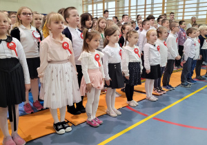 Duża grupa uczniów i uczennic odswiętnie ubranych stoi na sali podczas śpiewania hymnu Polski. Uczniowie mają przypięte biało-czerwone kotyliony na białych bluzeczkach