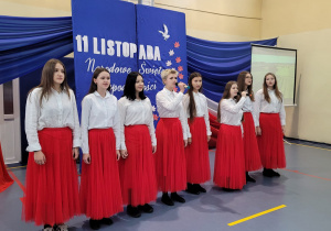 Grupa dziewcząt klas ósmych w biało czeronych strojach stoi na sali podczas śpiewania hymnu Polski