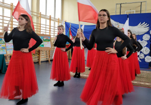 Grupa dziwecząt klas ósmych w czarnych bluzkach i czerwonych spódniczkach z flagami w dłoniach podczas tańca z flagami