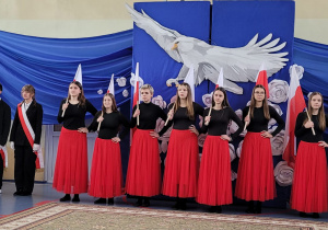 Grupa dziewcząt w czarno czerwonych strojach z flagami w rękach, Z lewej strony poczet sztandarowy szkoły.