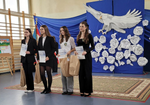 Cztery laureatki konkursu polonistycznego stoja na sali na tle dekoracji z Orłem Białym. Wsród uczennic Julia Bernackai Nikola Dębowska-Kuś