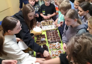 Duża grupa uczniów podczas kiermszu kupuje i degustuje ciasta.