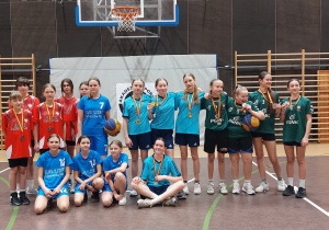 Cztery reprezentacje różnych szkół w strojach sportowych pozują na tle tablicy do kosza.
