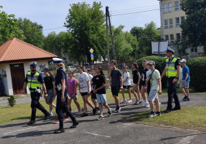 Grupa uczniów w eskorcie policji zmierza w kierunku placu manewrowego na egzamin z umiejetności jazdy na rowerzę i znajomości przepisów.