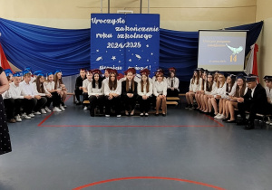 Wszyscy uczniowie klas ósmych siedzą na ławeczkach na tle dekoracji. Pani dyrektor przemawia.