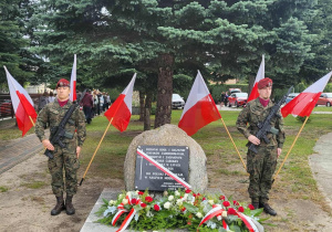Dwóch żołnierzy pełni wartę przy Pomniku Sybiraka w Tomaszowie Mazowieckim. Przed ponikiem wiązanki kwiatów, za żołnieżami flagi biało-czerwone.