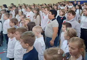Duża grupa uczniów szkoły podczas śpiewu hymnu państwowego.